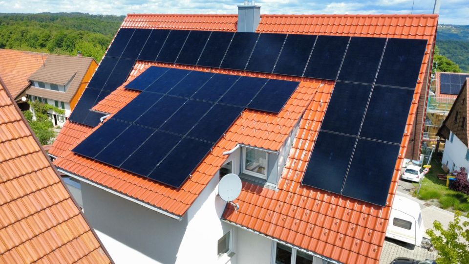 Schwarze Solarmodule auf einem roten Ziegeldach, im Hintergrund bewaldete Hügel